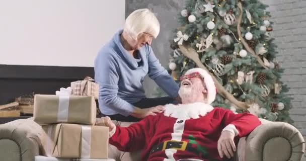 Kırmızı Noel şapkalı mutlu Kafkasyalı adam ve kanepeye oturmuş hediye kutularıyla gülümseyen sarışın kadın. Yaşlı çift yılbaşı ağacının önünde dinleniyor. Sinema 4k Prores Merkezi. — Stok video