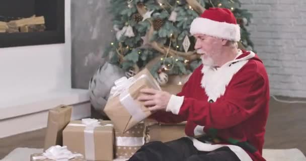 Viejo hombre caucásico con barba blanca y bigotes sosteniendo la caja de regalo y agitándola. Santa en sombrero rojo y suéter de Navidad sentado en el suelo bajo el árbol de Año Nuevo con regalos. Sede del cine 4k ProRes . — Vídeos de Stock