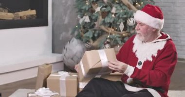 Noel Baba 'nın portresi Noel ağacının arka planında oturuyor ve hediye kutusunu sallıyor. Kırmızı şapkalı meraklı yaşlı adam yeni yıl arifesinde hediye seçiyor. Sinema 4k Prores Merkezi.