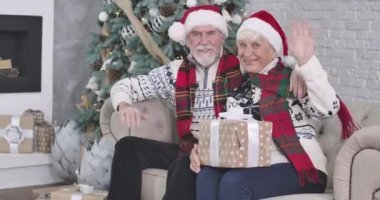 Orta boy, gülümseyen beyaz bir adam ve kadın kanepeye oturmuş elinde hediyelerle kameraya el sallıyor. Noel Baba ve Noel Anne, Noel arifesinde kameraya Mutlu Noeller diliyor. Sinema 4k Prores Merkezi