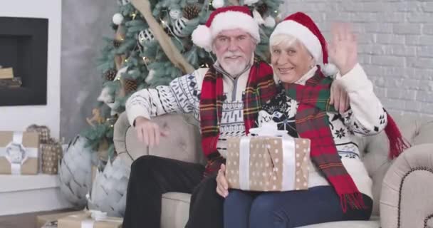 Közepes lövés, mosolygó öreg fehér férfi és nő ül a kanapén ajándékokkal és integet a kamerának. Boldog Mikulást és Mrs. Claus Boldog Karácsonyt kívánnak a kamera előtt szilveszterkor. Mozi 4k Prores Hq — Stock videók