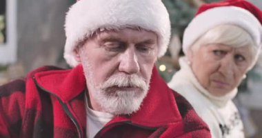 Kırmızı Noel şapkası takan, beyaz sakallı, üzgün, yaşlı, beyaz bir adamın yakın çekimi. Arka planda oturan üzgün kıdemli karısı, hoşnutsuz bir yüz ifadesiyle. Sinema 4k Prores Merkezi.