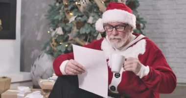 Noel Baba 'nın portresi kahve içerken, çay içerken ve mektup okurken. Gözlüklü, beyaz sakallı yaşlı adam yılbaşı arifesinde yazışmalara bakıyor. Sinema 4k Prores Merkezi.
