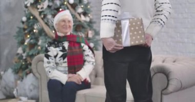 Beyaz bir adam arkasında hediye kutusu tutuyor. Gülümseyen ve alkışlayan yaşlı kadına odaklan. Koca, sevgili karısına hediye veriyor. Noel, yeni yıl. Sinema 4k Prores Merkezi..
