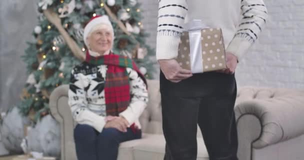 Hombre caucásico sosteniendo caja de regalo a sus espaldas. El enfoque cambia a mujer mayor sorprendida sonriendo y aplaudiendo. Esposo dando regalo a su encantadora esposa. Navidad, Año Nuevo. Sede del cine 4k ProRes .. — Vídeo de stock