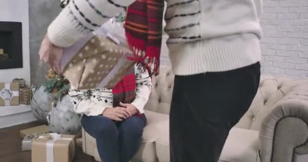 Überraschte kaukasische Seniorin, die auf der Couch neben dem Weihnachtsbaum saß, als ihr Mann ihr Geschenk machte und ihr auf die Wange küsste. Weihnachten, Neujahr, Feiertage, Überraschung. kino 4k prores hq. — Stockvideo