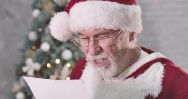 Noel Baba 'nın mektup okuyup suratını buruşturduğu yakın plan. Beyaz sakallı son sınıf öğrencisi, seçeceği hediyeyi düşünüyor. Noel ağacının arka planında yorgun Noel Baba. Sinema 4k Prores Merkezi.
