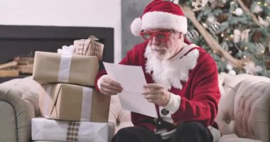 Noel Baba 'nın portresi hediye kutularıyla oturuyor ve mektupları okuyor. Kırmızı gözlüklü yaşlı adam yeni yıl dileklerini inceliyor. Arka planda duran Noel ağacı. Sinema 4k Prores Merkezi.
