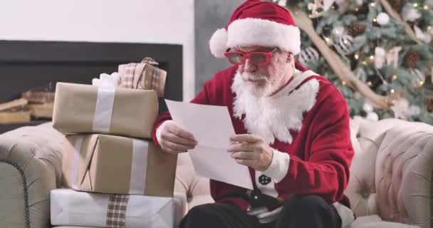 Porträtt av jultomten sitter med presentaskar och läsa brev. Gamling i röda glasögon som undersöker folks önskningar om nyår. Julgran står i bakgrunden. Cinema 4k Prores Hq. — Stockvideo