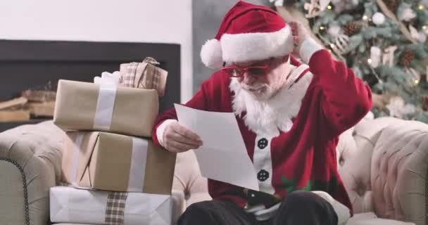 Összpontosított Mikulás ül kanapén előtt karácsonyfa és olvasás betűk. Érett férfi vörös szemüvegben, vakarja és rázza a fejét. Mikulás gondolkodik szilveszterkor. Mozi 4k Prores Hq.