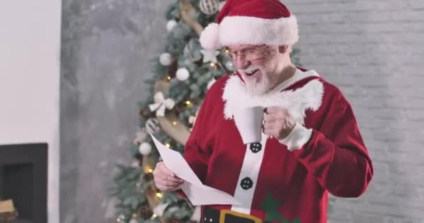 Smiling old Caucasian man in Santa Claus costume drinking coffee and reading letter. Santa in eyeglasses smiling as standing at the background of Christmas tree. Cinema 4k ProRes HQ. — Stock Video