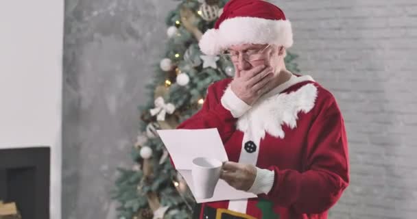 Thoughtful old Caucasian man in Santa Claus costume reading letter, stroking white beard and scratching head. Santa in eyeglasses examining correspondence on New Years eve. Cinema 4k ProRes HQ. — Stock Video