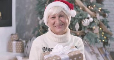 Son sınıf öğrencisi, elinde Noel hediyesi kutusuyla kameraya bakıp gülümseyen beyaz bir kadının yakın plan portresi. Güzel yaşlı bayan yeni yıl arifesini evde geçiriyor. Sinema 4k Prores Merkezi.
