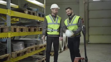 İki gülümseyen beyaz adam çelik metal deposunda dikilip konuşuyor. İşçiler üretim sahasında işlerini yapıyorlar. Üretim, üretim, meslek, meslek.