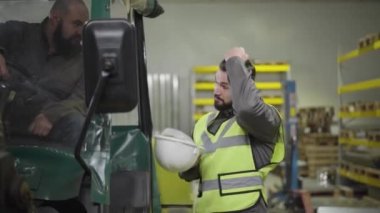 Genç sakallı beyaz adamın portresi kaskını takıyor ve traktördeki meslektaşıyla konuşuyor. Fabrikadaki işçiler. Üretim, üretim, profesyoneller. mesleği.