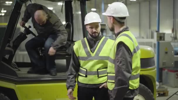 Retrato del hombre caucásico barbudo saliendo del tractor de remolque y saludando a sus colegas con cascos blancos y chalecos verdes. Trabajadores en planta de fabricación. Profesional, trabajador, cargador, empleado, molino . — Vídeo de stock