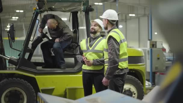 Jeunes hommes caucasiens debout à l'arrière-plan du tracteur de remorquage et parlant en tant que conducteur sortant de la cabine et serrant la main. Travailleurs dans l'usine de fabrication. Professionnel, travailleur, chargeur, employé — Video