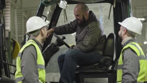 Two male Caucasian workers in helmets and vests talking to bearded colleague sitting in tow tractor. Plant employees working indoors. Manufacture, loader, production. — Stock Video