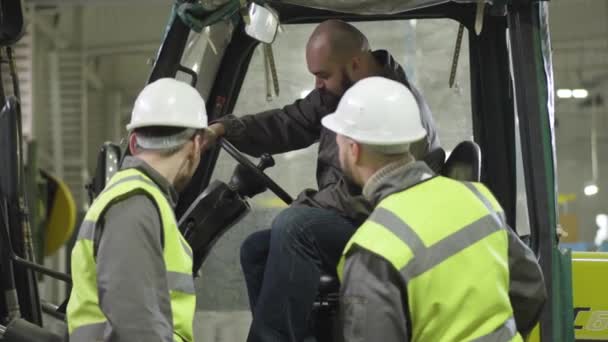 Portrait of bearded Caucasian tow tractor driver sitting in cabin and talking to his colleagues. Workers in vests and helmets having break at production site. Manufacture, loader, production. — Stock Video