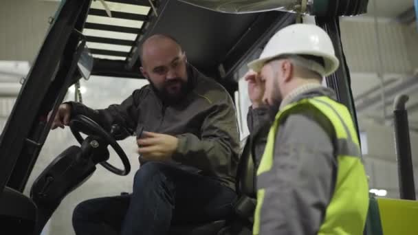 Portrait of bearded bald Caucasian man sitting in tow tractor cabin and talking to worker in helmet and vest. Employees having break at warehouse. Manufacture, working, warehouse, industry. — Stock Video