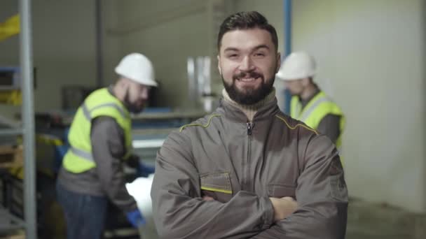 Portrét sebevědomého bělocha, který se dívá do kamery a usmívá se. Foreman s černými vlasy a vousy pózující na místě výroby na pozadí pracovníků. Výroba, mlýn, zaměstnanec. — Stock video