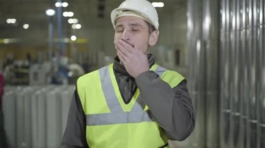 Kasklı ve yelekli yorgun beyaz adamın portresi çelik metal boruların arkasında esniyor ve geriniyor. Baca üretim sahasındaki işyerinde yorgun düşmüş bir işçi. Üretim, endüstri.