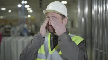 Miğfer ve yelek giymiş yorgun beyaz bir adam gözlerini ovuşturuyor ve üretim alanında geriniyor. Metal boruların arkasında duran yorgun genç işçi. Üretim, endüstri, çelik fabrikası.
