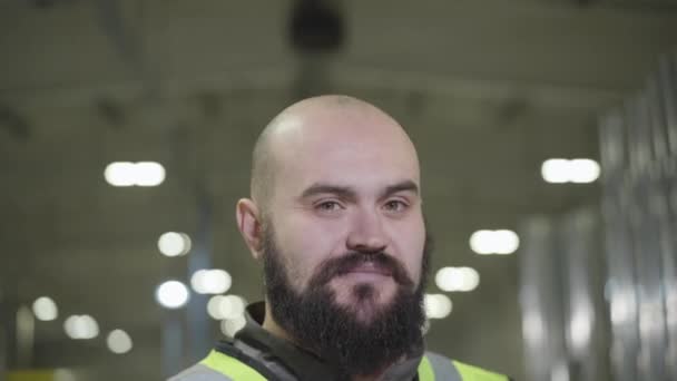 Cámara en movimiento hacia arriba, retrato de hombre caucásico calvo barbudo sonriendo a la cámara. Trabajador feliz en chaleco verde de pie en el sitio de producción. Planta, molino, fabricación, empleado, lugar de trabajo . — Vídeo de stock