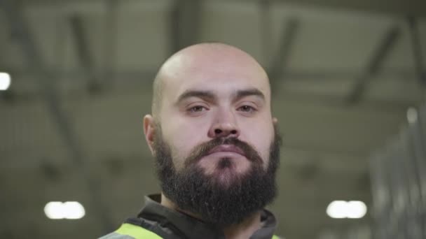 Närbild porträtt av allvarlig skallig kaukasisk man med svart skägg tittar på kameran. Anställd i grön väst stående vid produktionsanläggningen. Anläggning, fabrik, tillverkning, anställd, arbetsplats. — Stockvideo