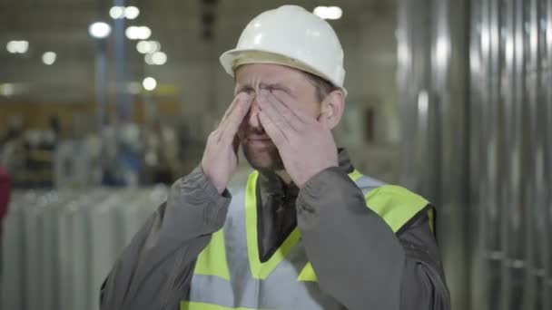 Homme caucasien épuisé en casque et gilet frottant les yeux et s'étirant sur le site de production. Fatigué jeune travailleur debout à l'arrière-plan de tuyaux métalliques en acier. Fabrication, industrie, aciérie .