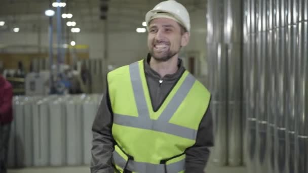 Porträt eines fröhlichen kaukasischen Arbeiters mit Schutzhelm, der vor dem Hintergrund von Stahlrohren tanzt. zufriedener Arbeiter an seinem Arbeitsplatz auf dem Schornsteinfertigungsgelände. Fertigung, Stahlindustrie. — Stockvideo