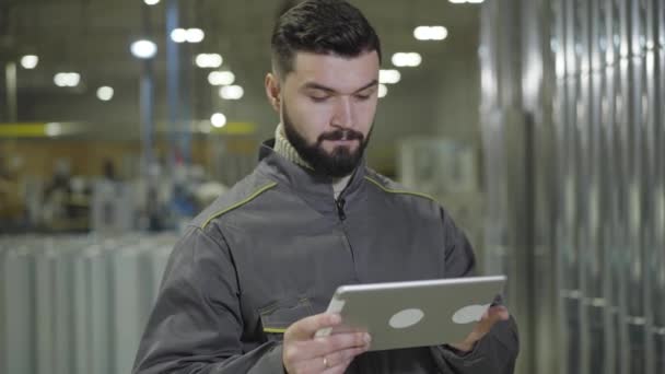 Porträtt av koncentrerad skäggig kaukasisk man som står vid produktionsstället och använder tabletter. Modern förman kollar stålrörslagret. Tillverkning, godsterminal, färdig produktion. — Stockvideo