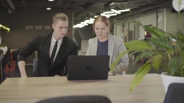 Portrait de jeune homme et femme caucasien en vêtements formels assis à la table dans un bureau à aire ouverte et à l'aide d'un ordinateur portable. Des collègues discutent du projet sur le lieu de travail. Entreprise, coworking . — Video