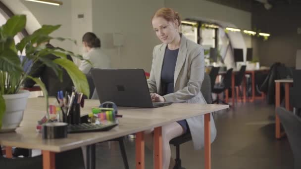 Retrato de mulher ruiva caucasiana confiante trabalhando no escritório usando laptop. Jovem trabalhadora de escritório em roupas formais elegantes que lidam com questões de negócios no local de trabalho. Coworking, espaço aberto . — Vídeo de Stock