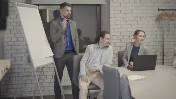 オープンスペースのオフィスでボードの横に立って、テーブルに座っている同僚と話を灰色のスーツに身を包んだ白人男性。ビジネスプロジェクトについて話し合う同僚。チームワーク、コーチング. — ストック動画