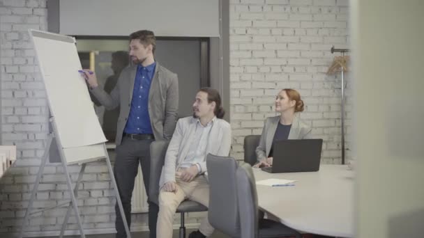 Homme caucasien adulte aux cheveux gris montrant des graphiques à bord dans un bureau ouvert à ses collègues. Des collègues assis à la table et discutant du projet. Travail d'équipe, coaching, direction, PDG . — Video