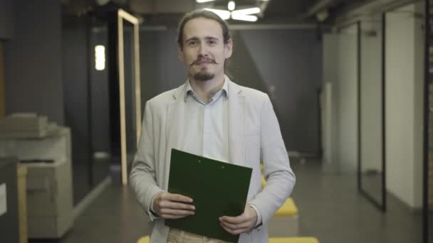 Retrato do jovem caucasiano com barba e bigodes olhando para a câmera e sorrindo. Gestor masculino em processo formal segurando pasta documento. CEO, gestão, empresário . — Vídeo de Stock