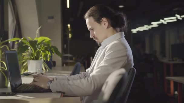 Kamera porusza się po skoncentrowanym białym mężczyźnie siedzącym w nocy w otwartej przestrzeni i wpisującym na klawiaturze laptopa. Portret Ceo w miejscu pracy. Biznes, sukces, przepracowanie, pracowitość. — Wideo stockowe