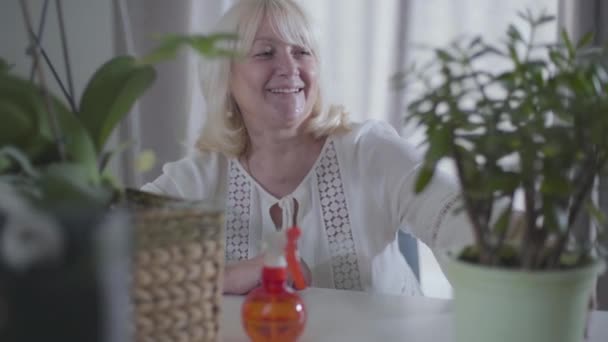 Glada blonda kaukasiska pensionär med trevligt leende och grå ögon tittar på hennes hem växter och ler. Porträtt av lycklig äldre kvinna som beundrar sin trädgård. hobby, tidsfördriv, fritid. — Stockvideo