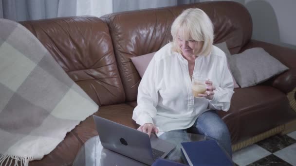 Visão superior da mulher caucasiana sênior atraente assistindo fotos ou fotos na tela do laptop e sorrindo. Loira madura aposentado usando laptop em casa. Atividades de lazer, passatempo . — Vídeo de Stock