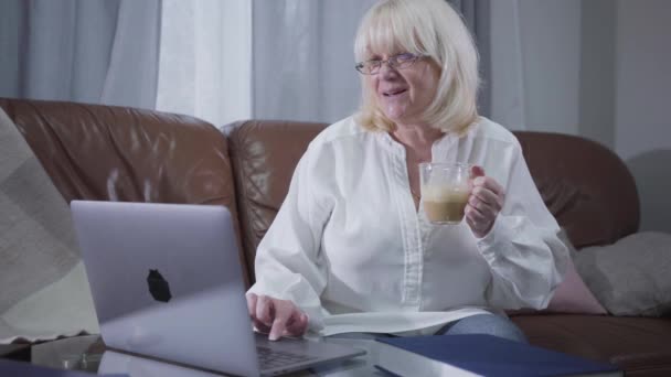 Mujer mayor caucásica positiva en anteojos sentados en el sofá con café y navegar por Internet. Rubia madura jubilada usando portátil en casa. Actividad recreativa, pasatiempo . — Vídeos de Stock