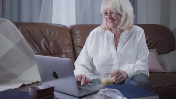 Πορτρέτο του Καυκάσιου συνταξιούχου fenmale σε γυαλιά που κάθεται στον καναπέ με φλιτζάνι καφέ και χρησιμοποιώντας φορητό υπολογιστή. Ξανθιά λευκή ώριμη γυναίκα σερφάρει στο διαδίκτυο στο σπίτι. Ψυχαγωγική δραστηριότητα, χόμπι. — Αρχείο Βίντεο