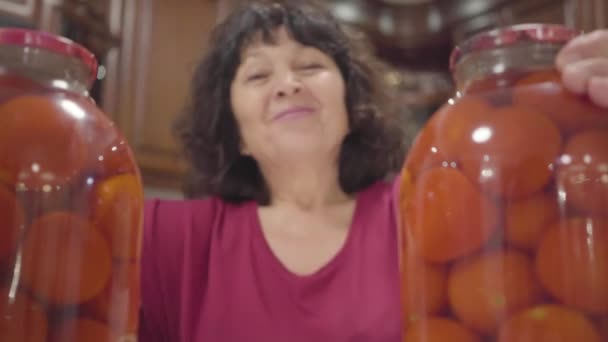 Mujer caucásica madura moviendo dos botellas aparte y sonriendo entre ellas. Mujer positiva jubilada admirando sus tomates enlatados. Feliz abuela preparando verduras de temporada en casa . — Vídeo de stock