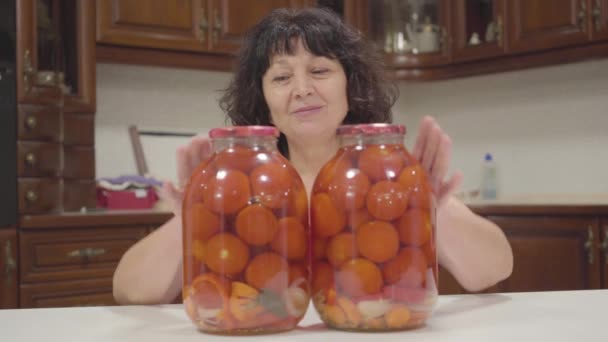 Pozytywna biała kobieta rozsuwa dwie butelki i pokazuje kciuki na obu dłoniach. Pozytywna emerytka pokazująca swoje puszkowane pomidory i uśmiechnięta. Szczęśliwa babcia przygotowuje sezonowe warzywa. — Wideo stockowe