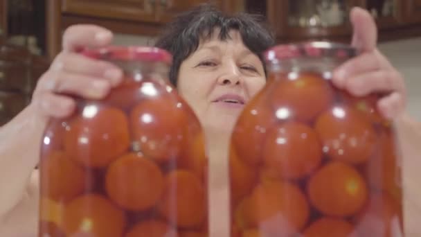 Cara de cerca de la feliz jubilada caucásica moviendo botellas con tomates enlatados separados, mostrando el pulgar hacia arriba y guiñando un ojo. Mujer mayor orgullosa de las verduras enlatadas. Abuela preparando comida de temporada . — Vídeo de stock