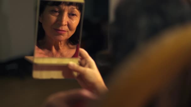 Close-up van kleine spiegel met reflectie van senior blanke vrouw die haar krullend haar repareert en praat. Positieve vrouwelijke gepensioneerde die 's avonds thuis rust. gepensioneerd herinnerend aan haar schoonheid en jeugd. — Stockvideo