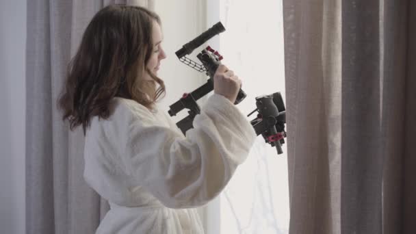 Retrato de vista lateral de una joven morena caucásica parada junto a la ventana con equipo de video. Hermosa camarógrafa sosteniendo el estabilizador por la mañana. Profesión, creatividad, estilo de vida . — Vídeos de Stock