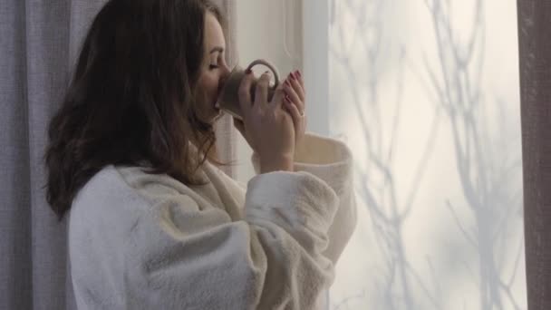 Vista lateral close-up da jovem bela mulher bebendo café na janela e fixando roupão branco. Menina branca sorridente desfrutando de manhã em casa. Lazer, estilo de vida, fins de semana, férias . — Vídeo de Stock