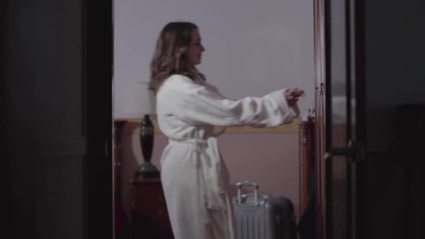 Shooting through doorway of beautiful young woman coming up to wardrobe, opening it, and looking inside. Caucasian girl in white bathrobe choosing outfit in the morning. Comfort, coziness, lifestyle. — Stock Video