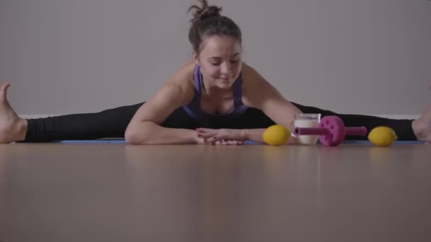 Portret van een jong, sportief meisje, zittend op het touw en buigend op de vloer. Rol wiel en sinaasappels liggen naast blanke brunette vrouw. Gezonde levensstijl, workout, sporten. — Stockvideo